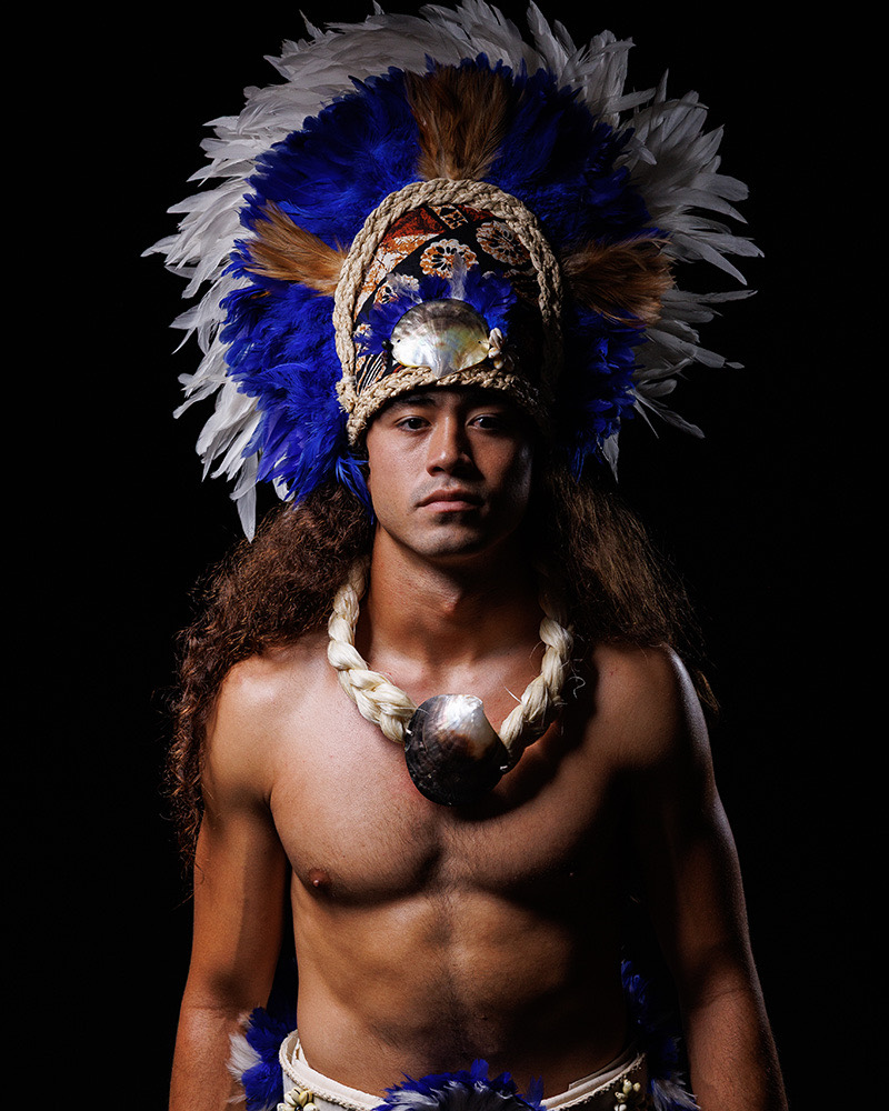 Male performer in a blue and white feathered headdress and a braided cord necklace with a shell pendant, against a black background