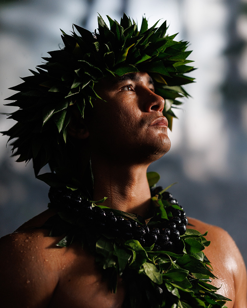 Man wearing a green leaf head lei and a dark bead lei, looking upward into soft light