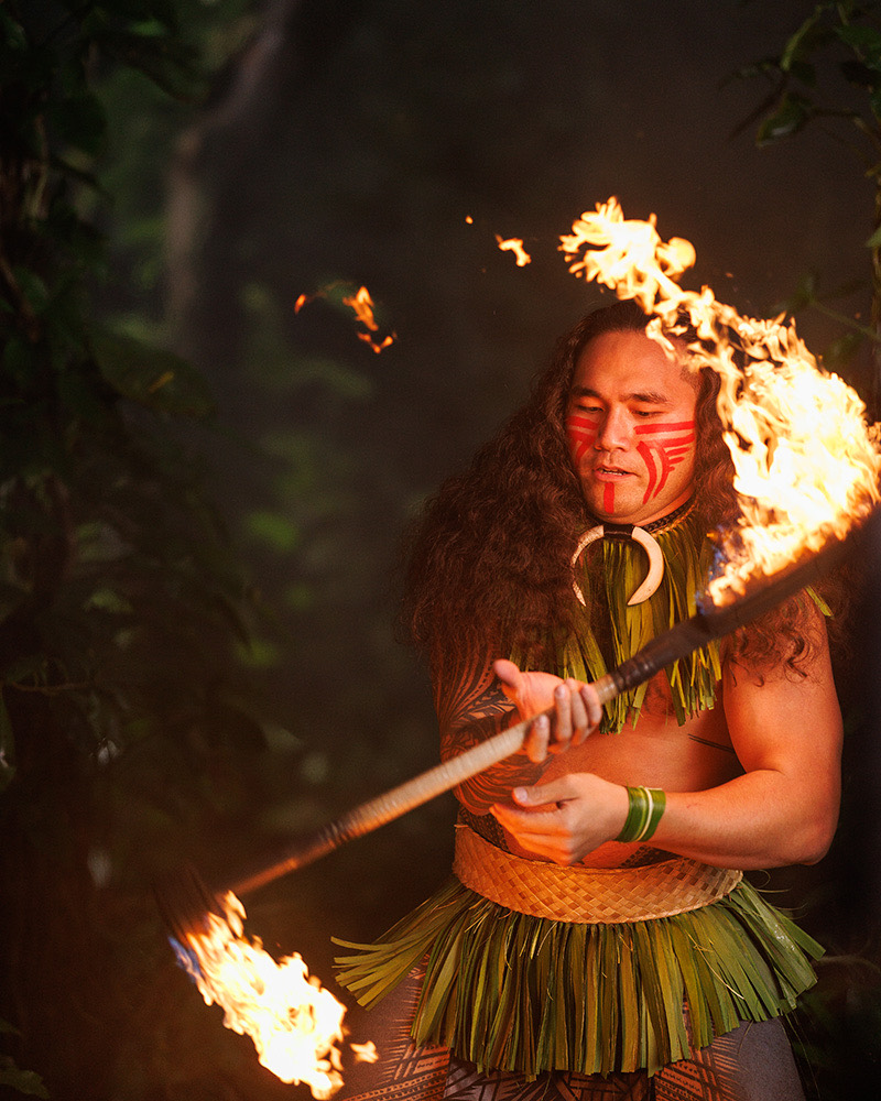 Fire knife performer in a leaf skirt and red face paint spinning a flaming fire knife in a dark forest