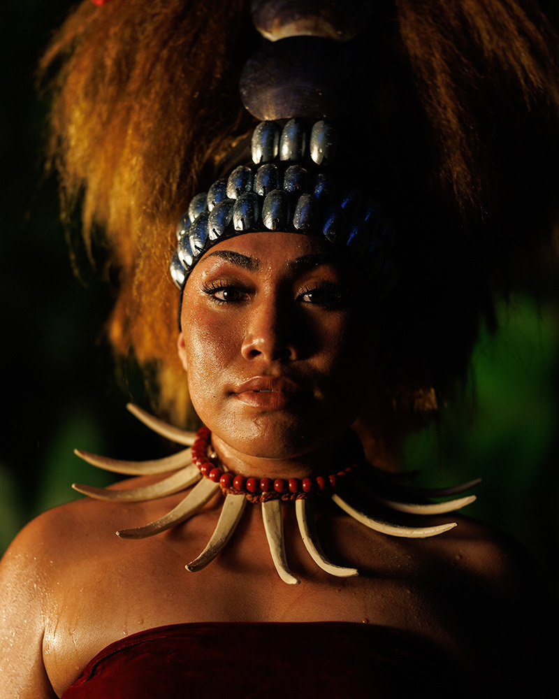 Portrait of a woman wearing a beaded headband, a red bead necklace and a tusk collar, lit warmly against dark foliage