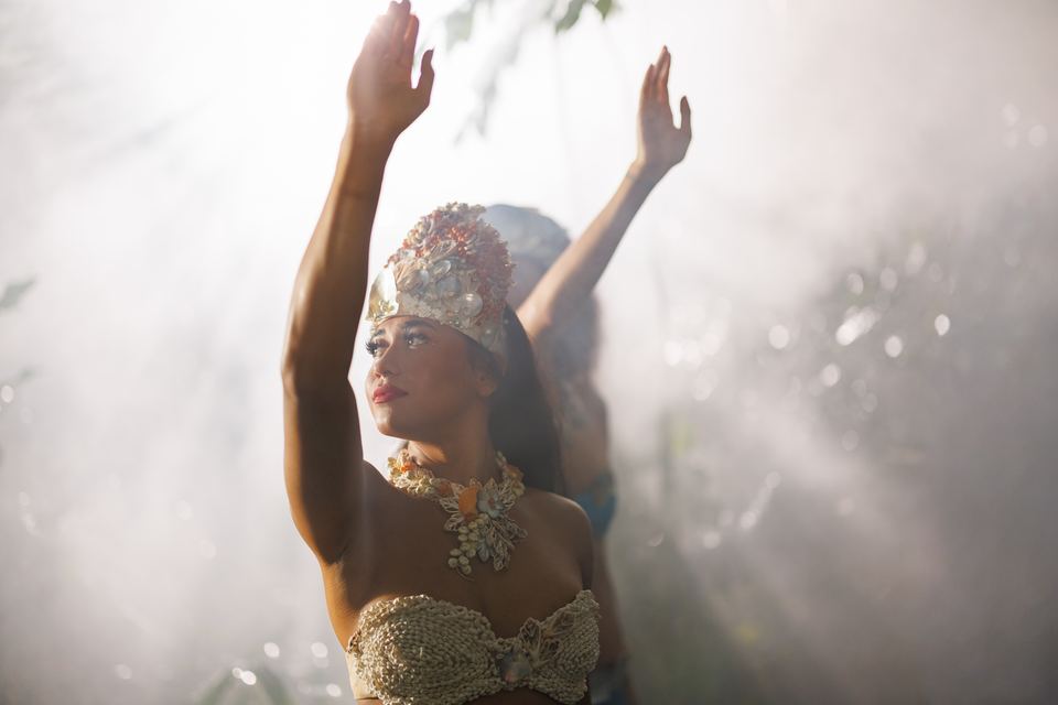 Dancer in a shell crown and shell top raises both arms, a second dancer behind her, as sunlight streams through white forest mist.