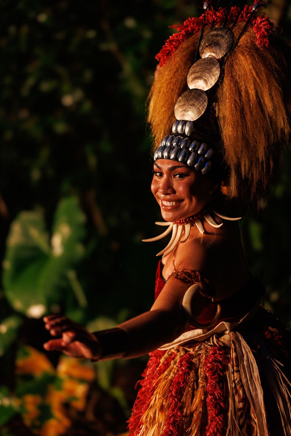 Smiling dancer in a Samoan tuiga headdress and tusk necklace looks back over her shoulder among tropical leaves.