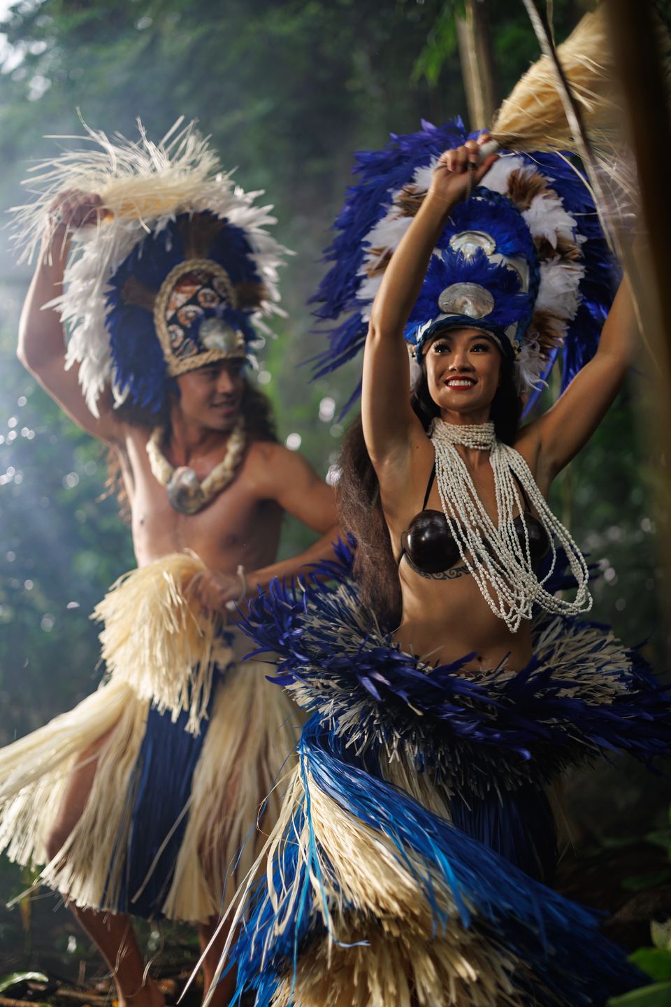 Two dancers in blue and white feather headdresses and raffia skirts raise their arms mid-dance in a misty forest.