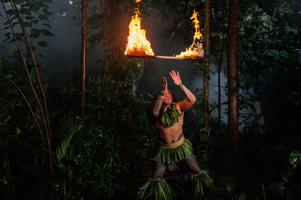 Fire knife dancer with red face paint and a green ti-leaf skirt reaches up toward a knife flaming at both ends above him in a smoky forest.