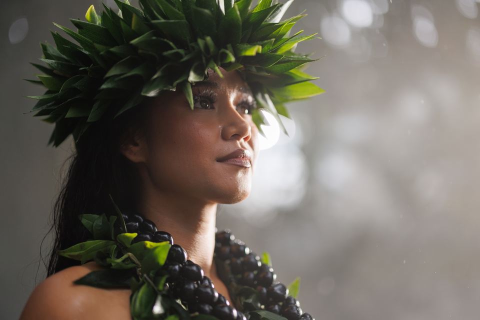 Hula dancer in a leaf crown and black kukui nut lei smiles slightly, looking off-frame against soft rain-speckled light.