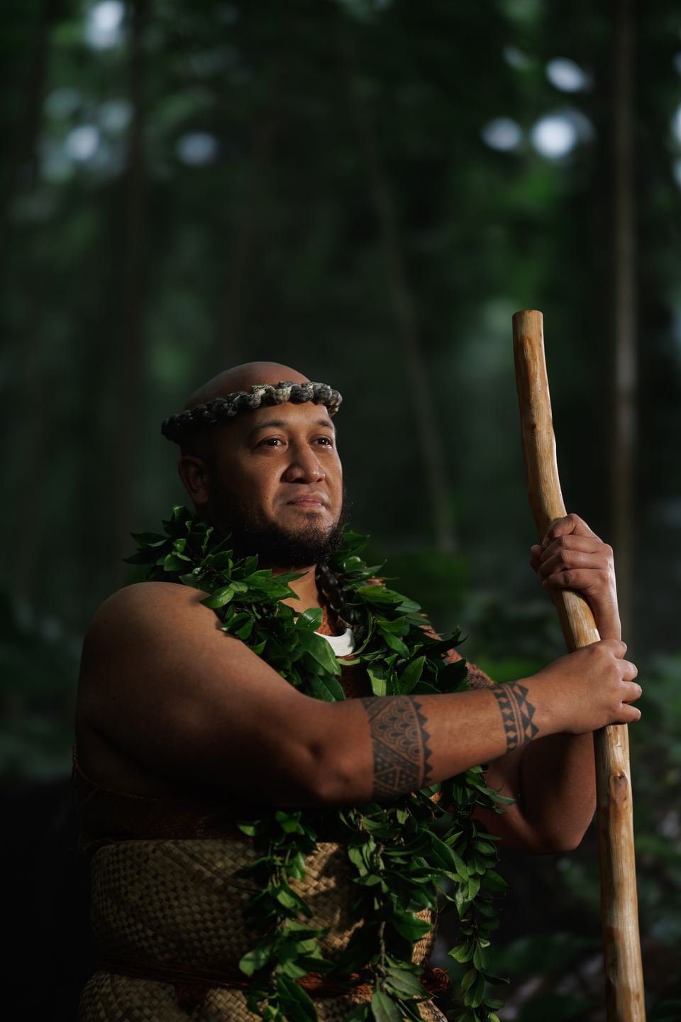 Man in a shell headband and leaf lei, with tattooed forearms, rests both hands on a wooden staff in a green forest.