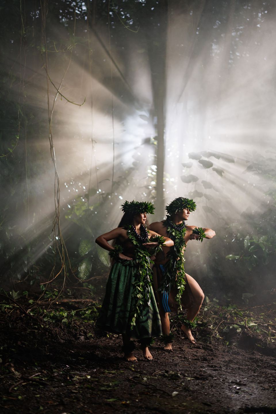 Two hula dancers in leaf crowns step in profile on a forest floor as sunbeams stream through the mist.