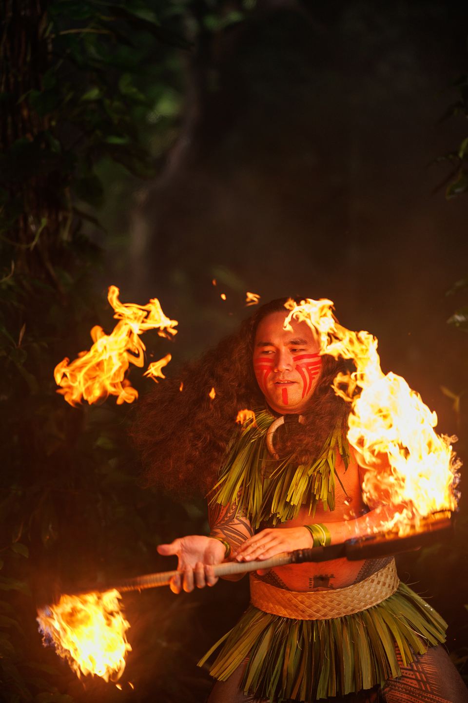 Fire knife dancer with red face paint twirls a knife flaming at both ends, the fire framing his face.