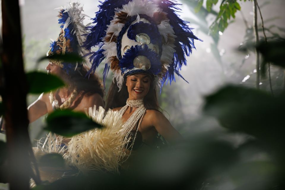 Seen through blurred leaves, a smiling dancer in a blue and white feather headdress moves beside her partner in the mist.
