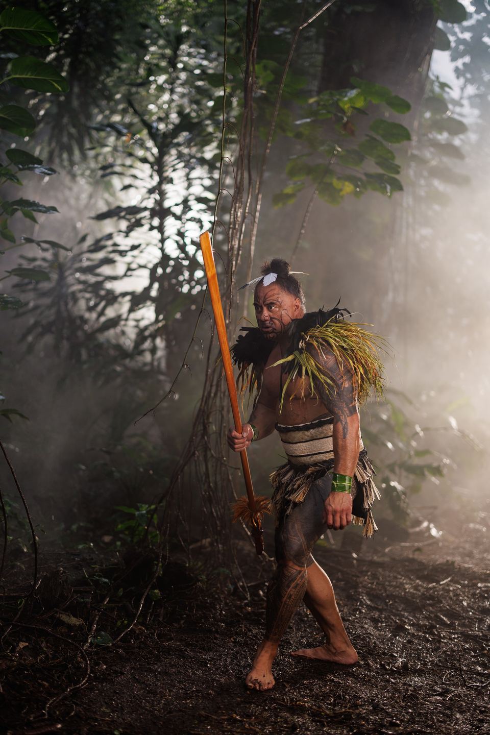 Māori performer with tā moko and a feather and flax shoulder cape strides barefoot through a misty forest carrying a wooden taiaha.