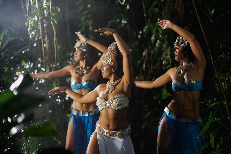Three dancers in shell crowns, one in white and two in blue skirts, dance with curved arms in a dark rainforest with drifting mist.