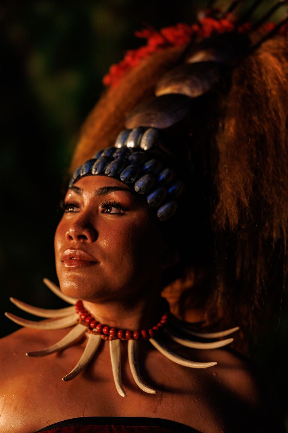 Woman in a Samoan tuiga headdress of hair, shells and red feathers, with a tusk necklace, gazes upward in warm light.