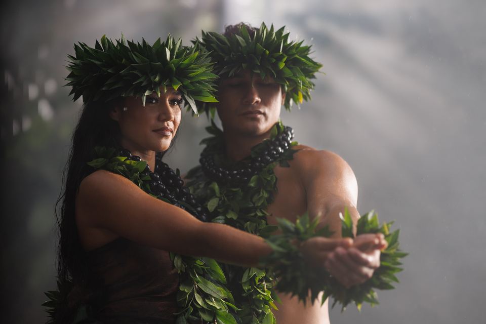 Two hula dancers in leaf crowns and kukui nut lei extend cupped hands together in soft, hazy light.