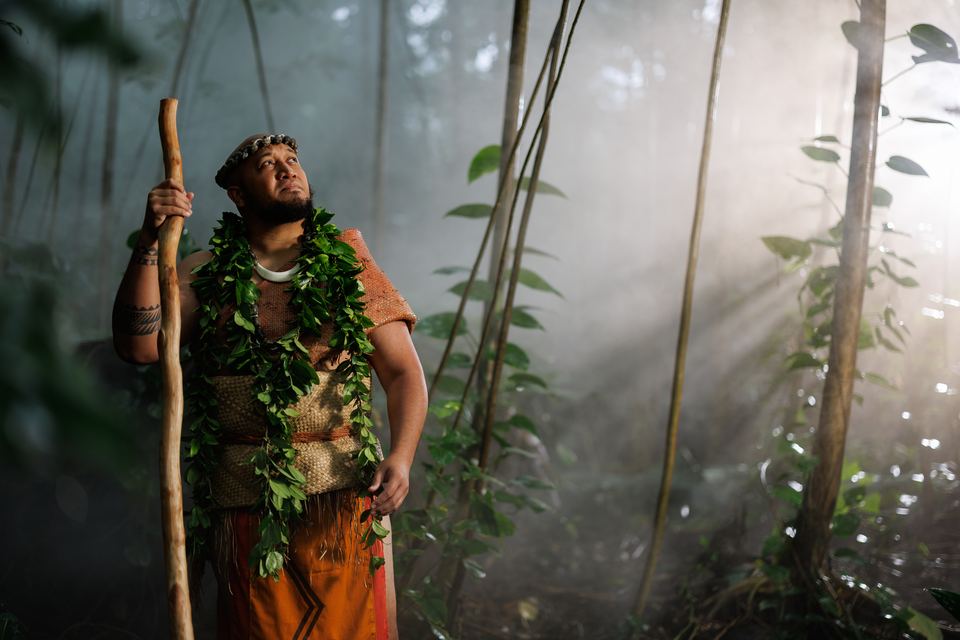 Man in a leaf lei, woven vest and orange wrap holds a tall wooden staff as sun rays cut through forest mist.