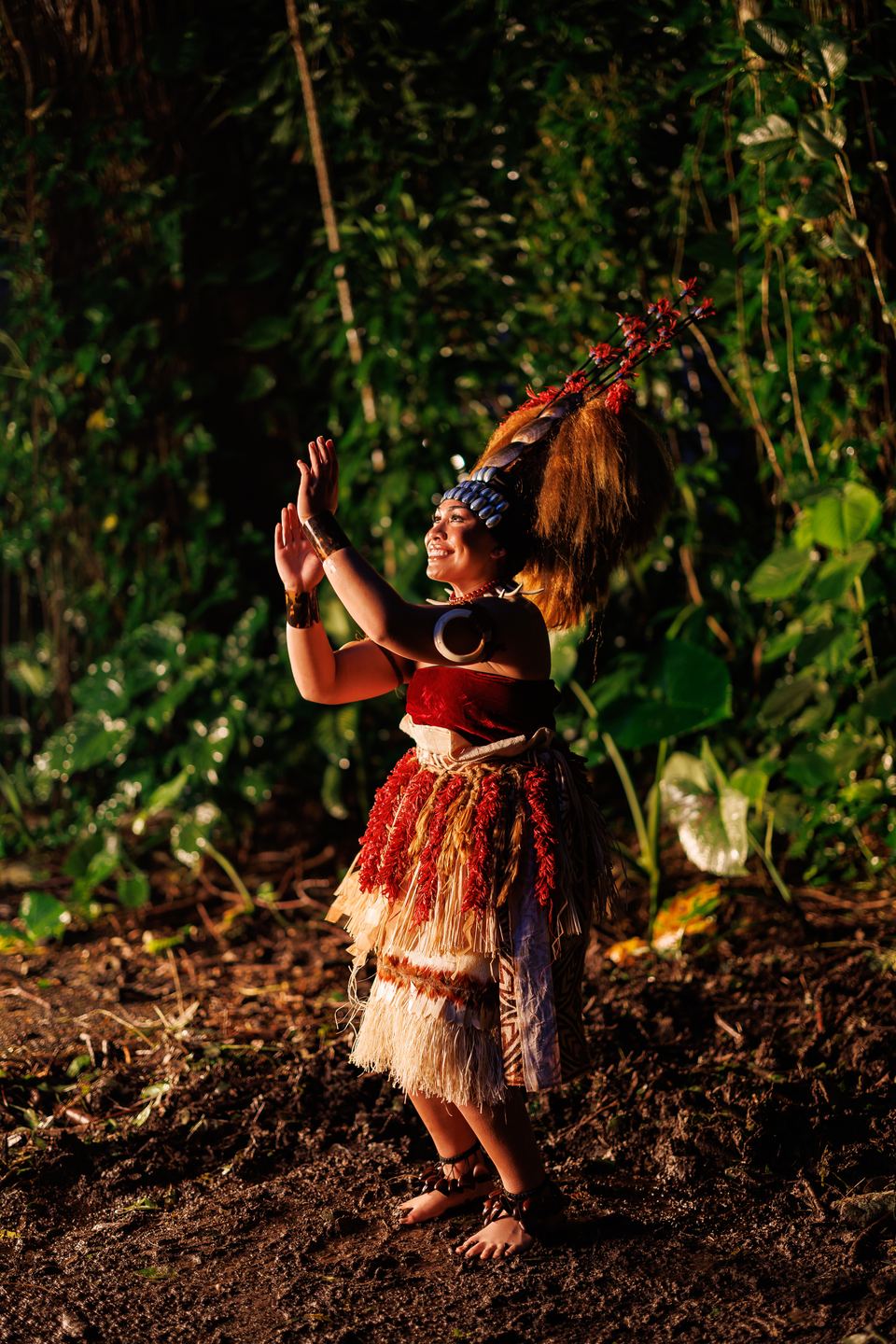 Dancer in a Samoan tuiga headdress and red-fringed fiber skirt claps with raised hands on a sunlit forest floor.