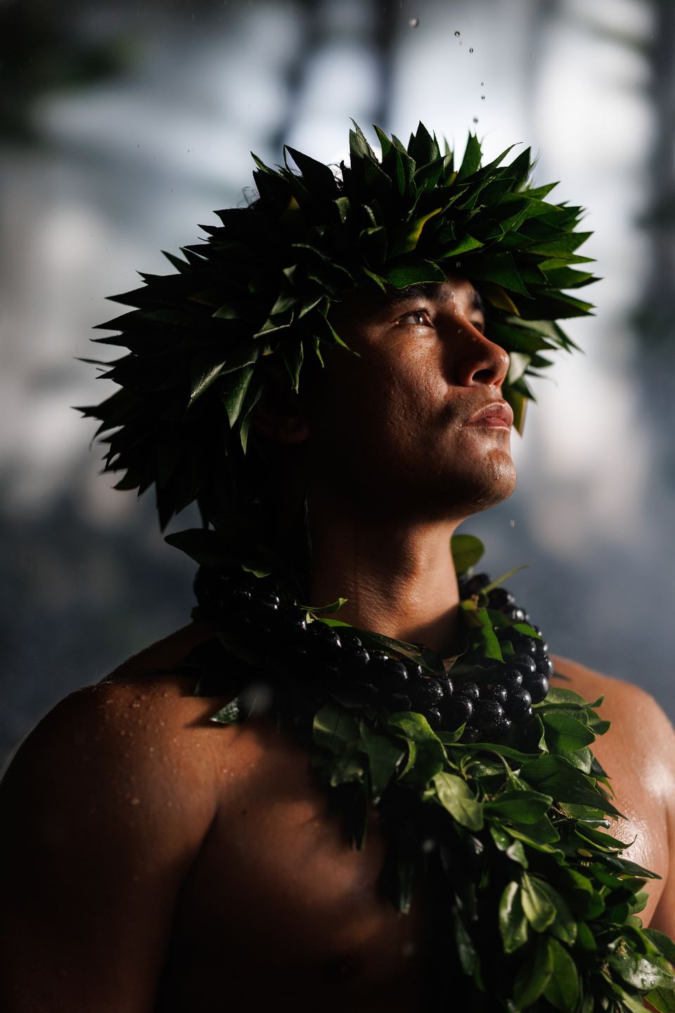 Male hula dancer in a leaf crown and kukui nut lei gazes upward as water droplets fall in soft backlight.