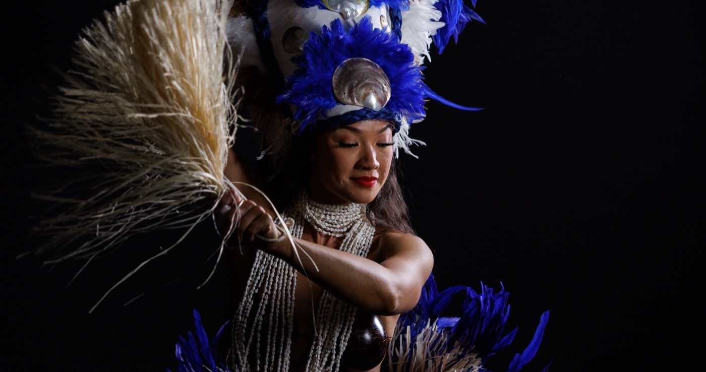 Dancer in a blue and white feathered headdress with pearl-shell adornments, holding a raffia fan against a dark backdrop