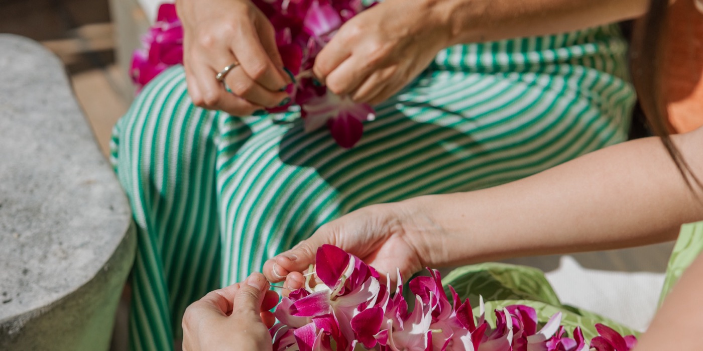 Two people stringing pink orchid blossoms into lei