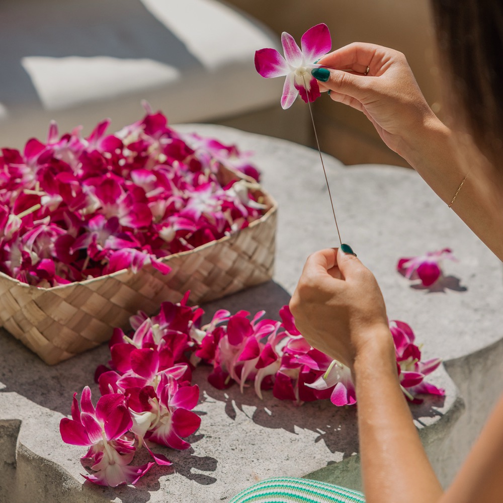 Hands stringing pink orchid blossoms into a lei beside a woven basket of orchids
